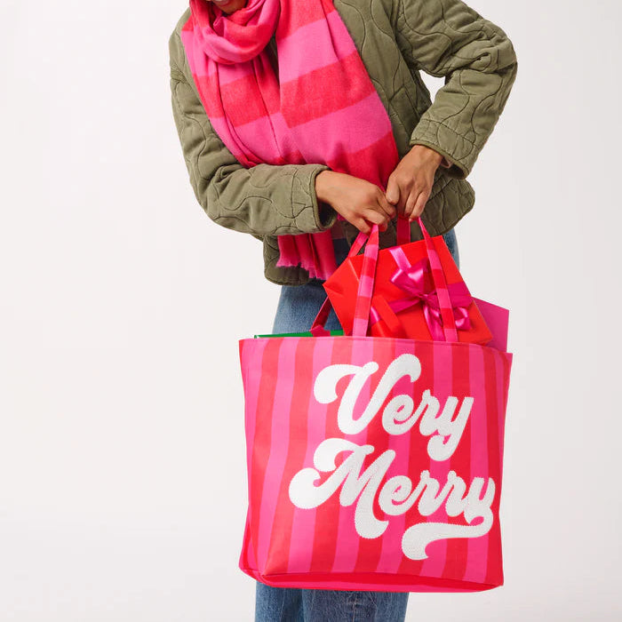 Photograph of a person in a studio setting wearing an olive green quilted jacket and striped scarf while carrying a large pink and red striped tote bag with Very Merry printed in white script and a red gift box with a pink bow