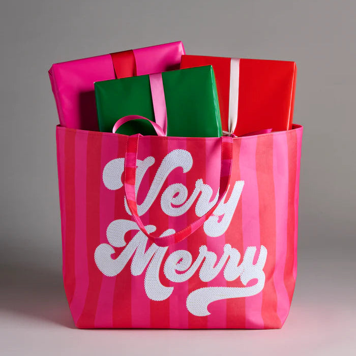 Studio photograph of a pink and red striped tote bag featuring white Very Merry script in a neutral studio containing glossy wrapped gifts in vibrant fuchsia, emerald green, and crimson red with satin ribbons