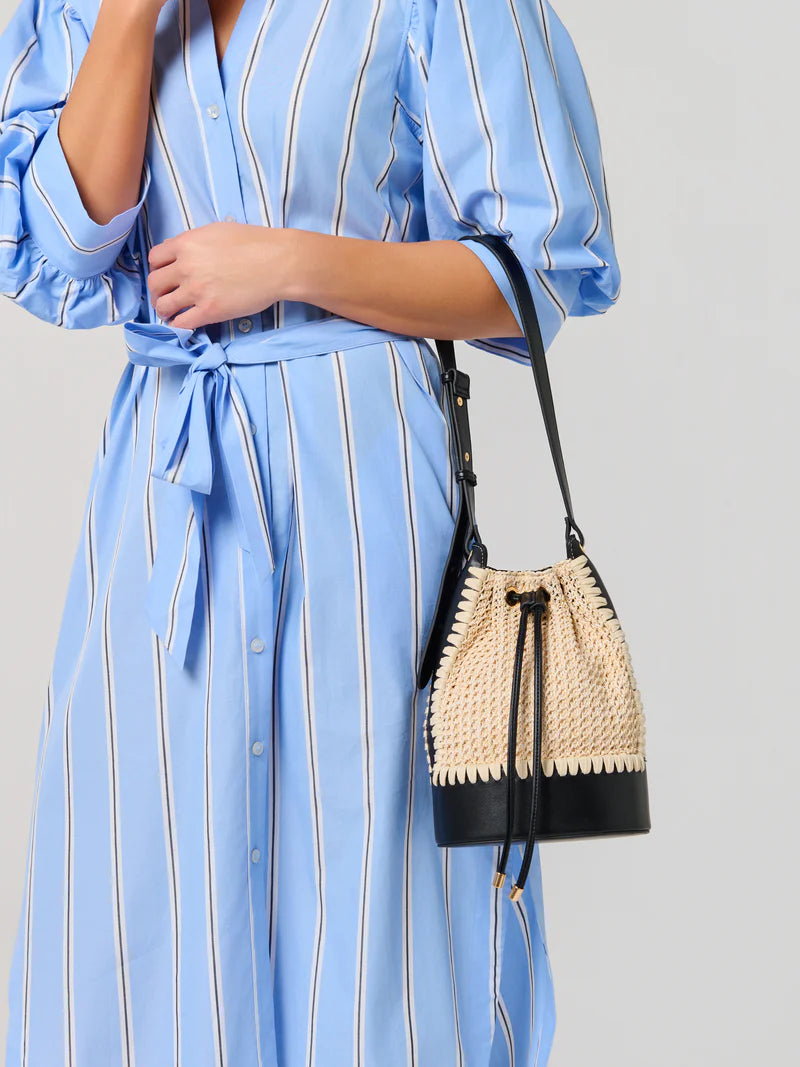 Photograph of a woman in a studio setting wearing a light blue and white striped dress while carrying a woven straw bucket bag featuring a black leather base and gold-tipped drawstrings