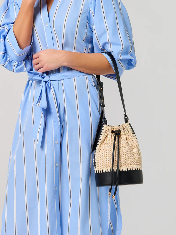 Photograph of a woman in a studio setting wearing a light blue and white striped dress while carrying a woven straw bucket bag featuring a black leather base and gold-tipped drawstrings