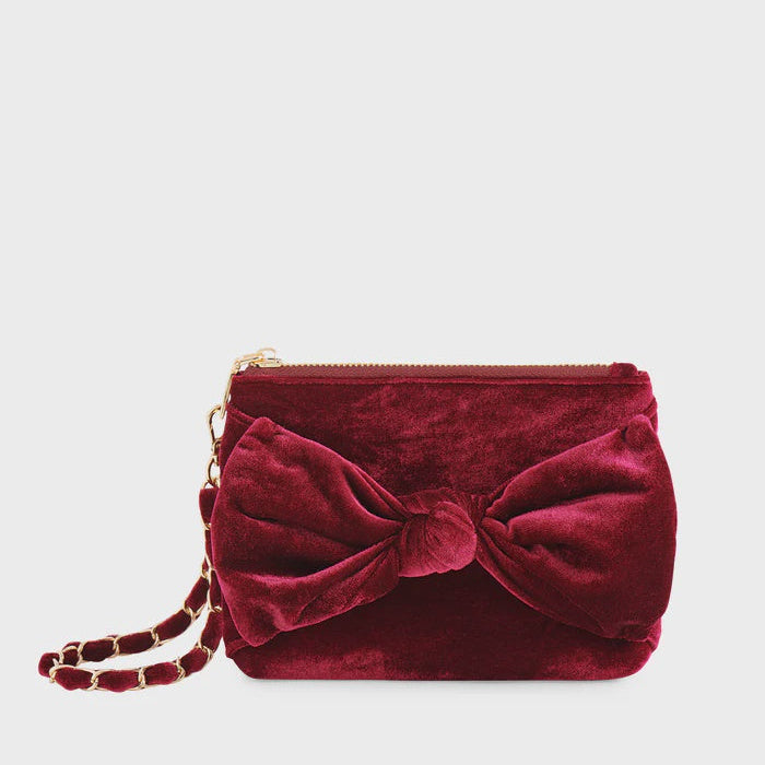 Velvet textile fashion accessory burgundy clutch handbag positioned against a neutral studio backdrop featuring a large decorative knotted bow and a gold-finish chain strap interwoven with matching fabric