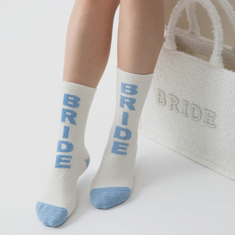 Product photograph of a person wearing fuzzy white socks with vertical light blue BRIDE lettering and blue-tipped toes positioned next to a matching textured white tote bag in a minimalist studio setting