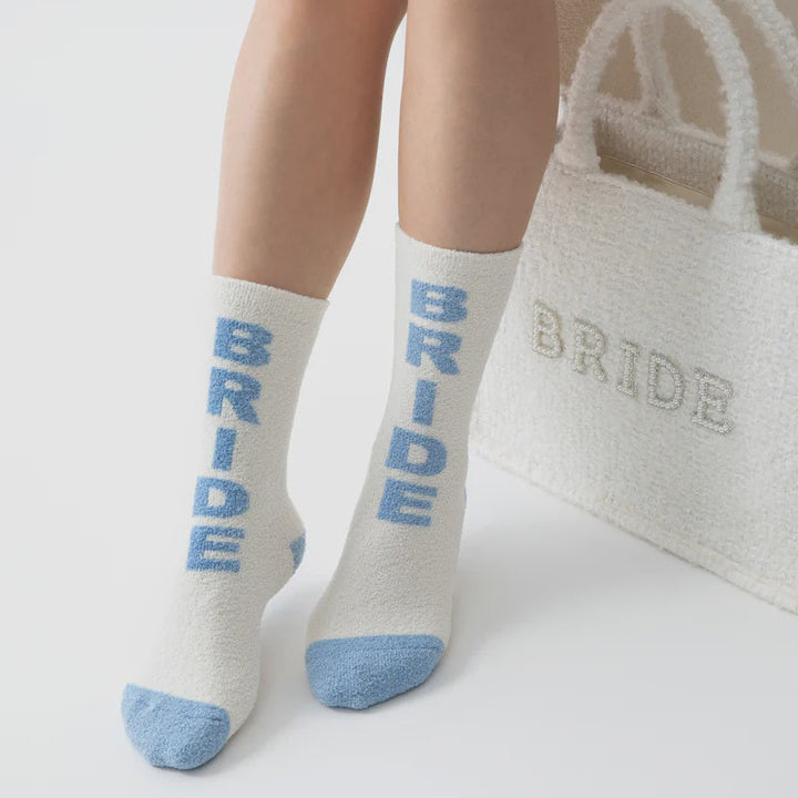 Product photograph of a person wearing fuzzy white socks with vertical light blue BRIDE lettering and blue-tipped toes positioned next to a matching textured white tote bag in a minimalist studio setting