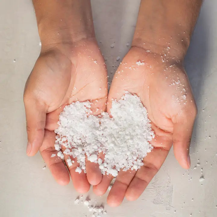 Photograph of a person's cupped hands holding a heart-shaped mound of coarse white sea salt over a neutral background with scattered translucent crystals visible on the palms and fingertips