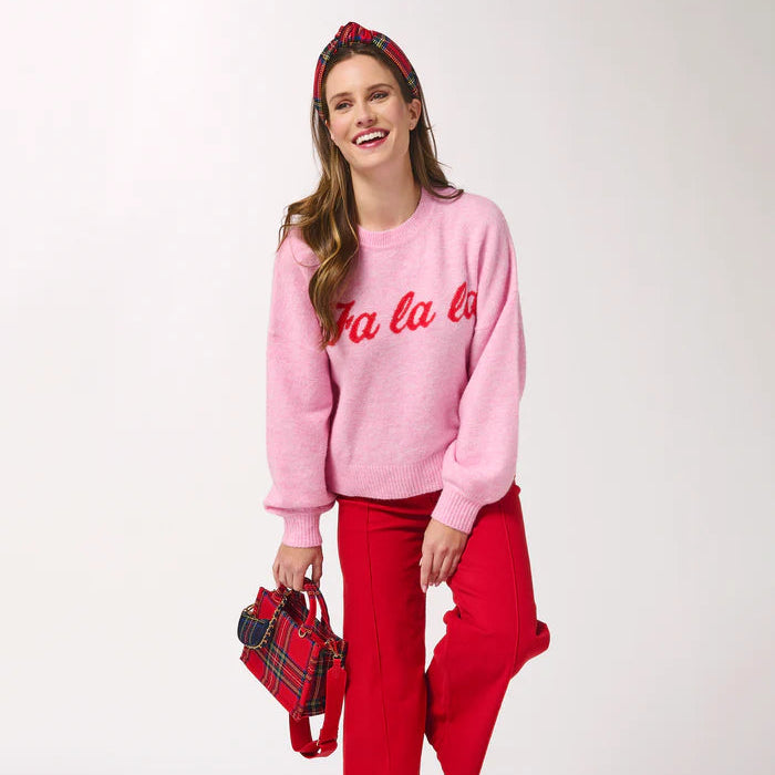 Studio photograph of a woman posing against a white backdrop wearing a pink holiday sweater featuring red fa la la script, bright red trousers, and matching tartan plaid accessories including a knotted headband and small handbag