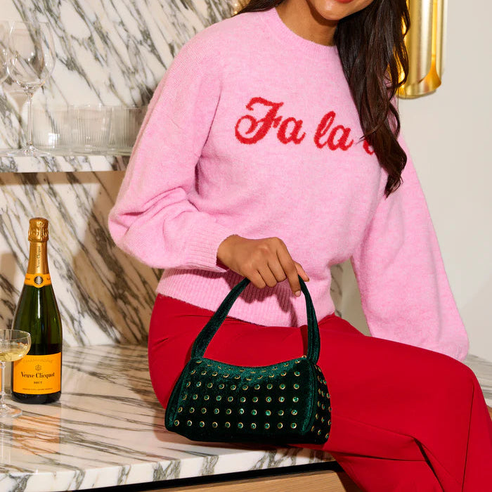 Digital lifestyle photograph of a woman wearing a pink knit Fa la sweater and red trousers seated on a white marble counter holding a studded emerald green velvet handbag beside a bottle of Veuve Clicquot champagne