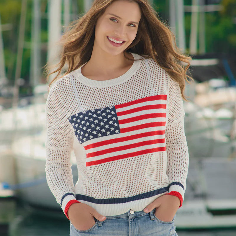 Digital photograph of a smiling woman at a coastal marina wearing a white mesh knit sweater with an American flag graphic, featuring red and blue striped cuffs and light-wash jeans with blurred sailboat masts in the background