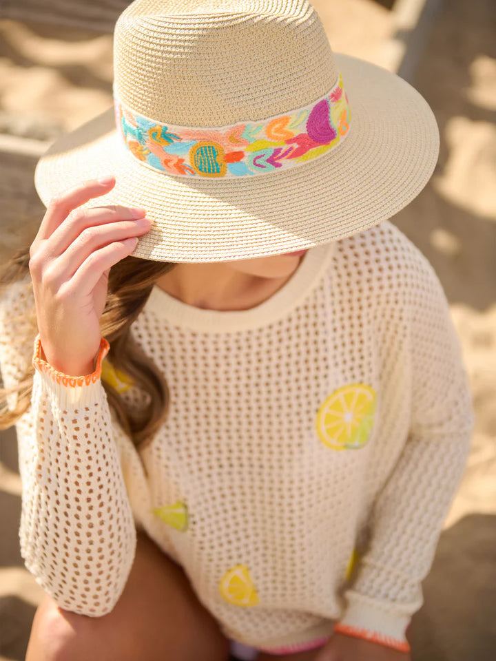 Photograph of a woman on a sandy beach wearing a woven straw hat with a colorful embroidered band and a white mesh sweater featuring bright yellow lemon slice patches
