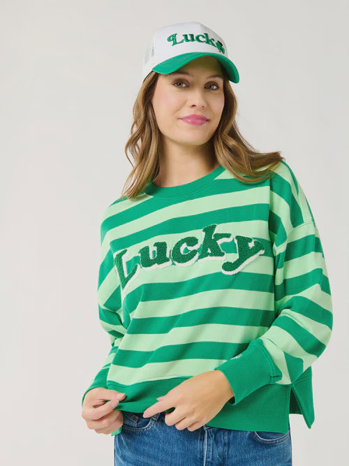 Digital photograph of a woman posing in a studio wearing a green striped St. Patrick's Day sweatshirt with a textured chenille Lucky patch and a matching white and green trucker hat featuring an embroidered shamrock logo