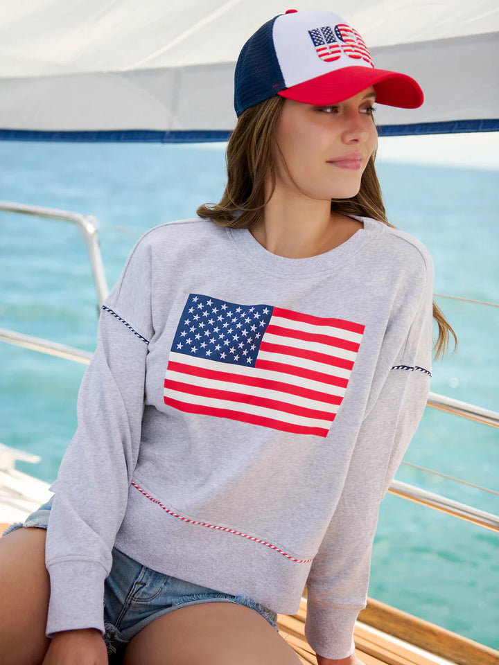 Lifestyle photograph of a woman on a boat wearing a light gray sweatshirt with a bold American flag graphic and red-and-white braided trim, a red and navy blue trucker hat with a patriotic emblem, and denim shorts against a backdrop of turquoise ocean water and silver boat railings