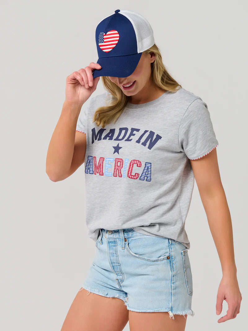 A digital studio photograph of a woman wearing a grey t-shirt with stylized Made In America lettering, frayed denim shorts, and a navy trucker cap featuring a heart-shaped American flag patch against a neutral background