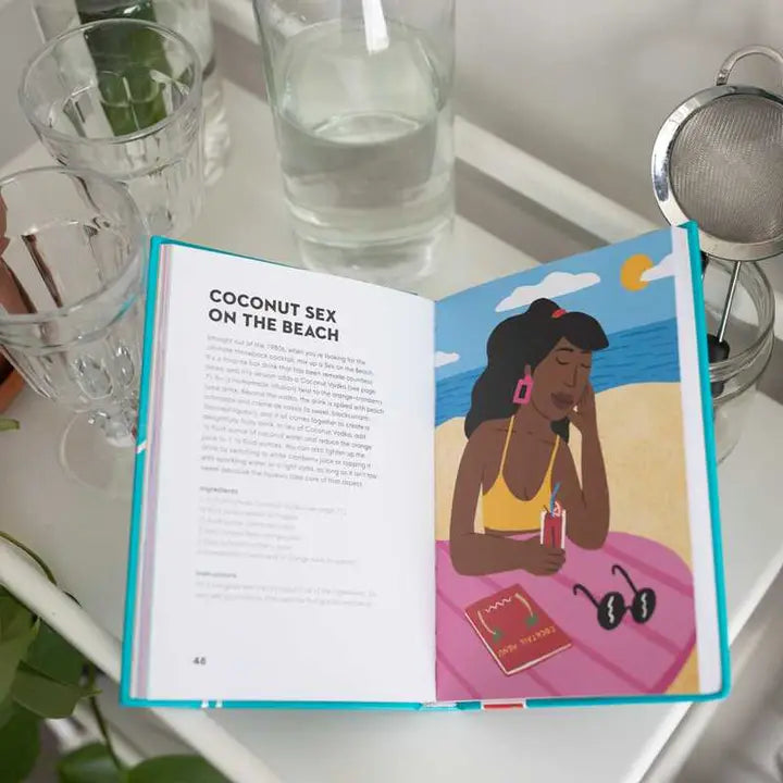 Photograph of an open mixology book featuring a vibrant illustration of a woman wearing pink rectangular earrings and a yellow bikini on a sunny beach, positioned next to fluted glassware and a silver cocktail strainer on a white surface