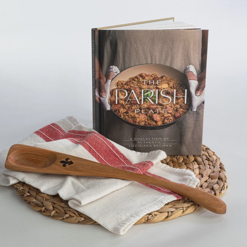 Studio photograph of The Parish Plate Louisiana cookbook standing on a woven seagrass mat beside a hand-carved wooden roux spoon with a burnt fleur-de-lis and a red-striped kitchen towel