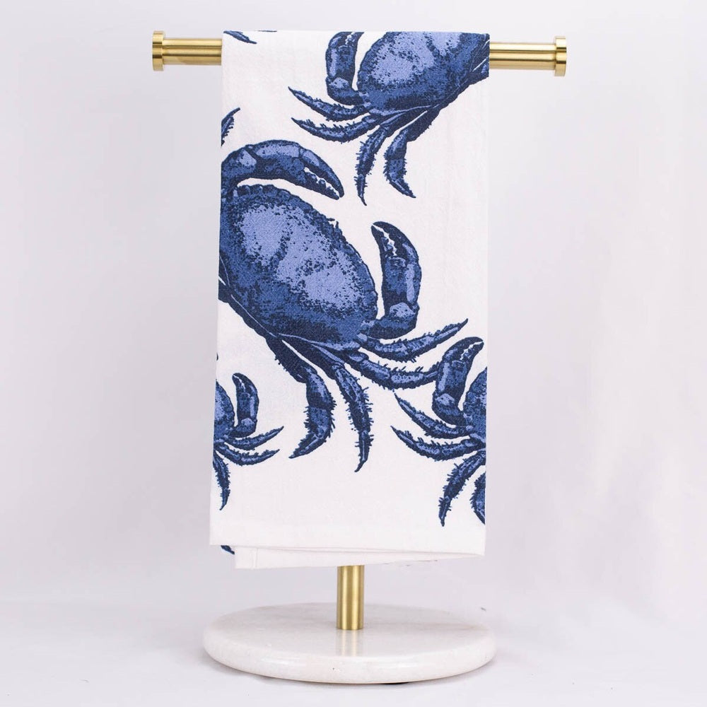 Printed cotton towel featuring detailed cobalt blue Maryland blue crab illustrations hanging on a modern gold metal T-bar rack with a circular white marble pedestal base