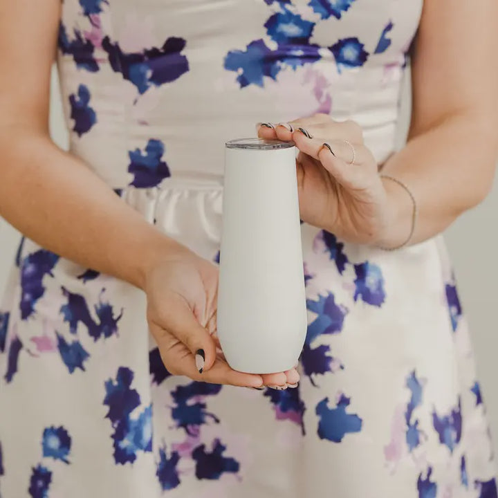 Photograph of a woman in an indoor setting holding a matte white insulated champagne flute while wearing a blue and purple floral dress and a black-tipped French manicure