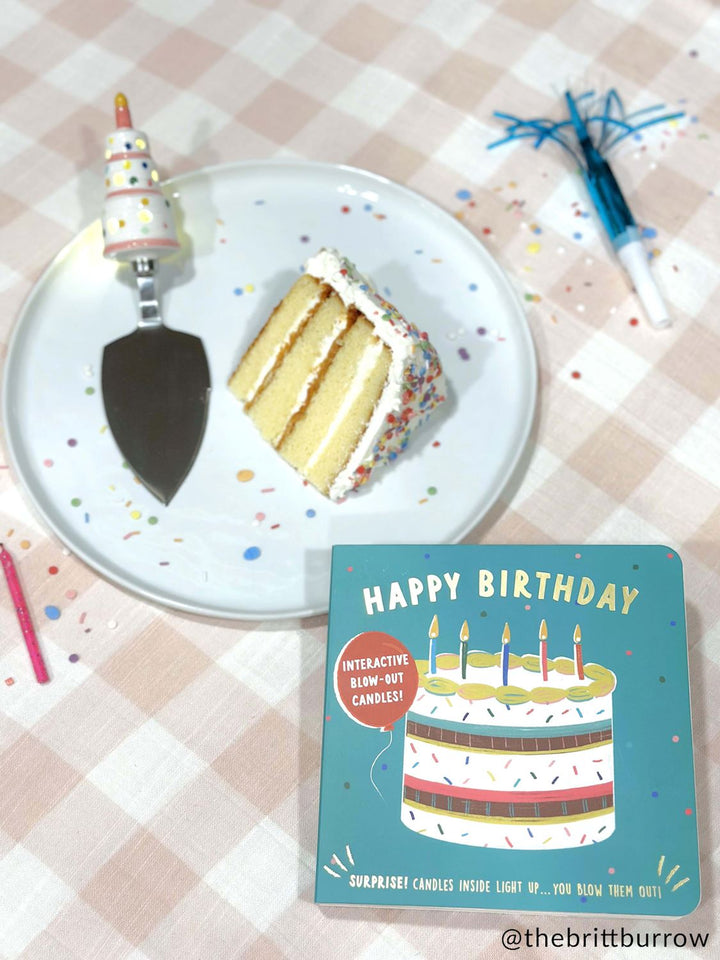 Photograph by @thebrittburrow of a slice of layered confetti cake on a white plate resting on a pink gingham tablecloth alongside a teal interactive Happy Birthday board book, a blue party blower, and a whimsical tiered-cake handle server