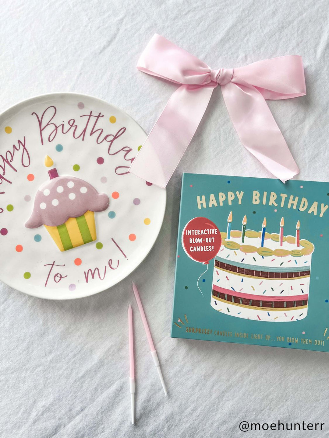 Photograph by @moehunterr of a birthday flat-lay on white fabric featuring a ceramic plate with a raised purple cupcake, a teal interactive book with a colorful cake illustration, and a large light pink satin ribbon bow