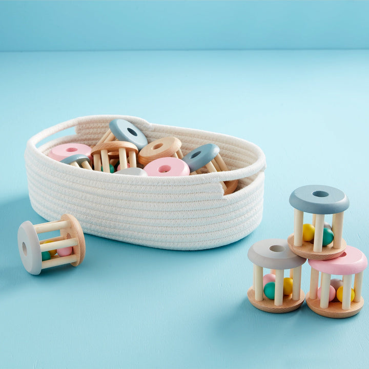 Studio photograph of wooden baby rattles featuring colorful interior rolling beads, with three toys stacked in a pyramid and several more resting in a white rope basket over a soft blue surface