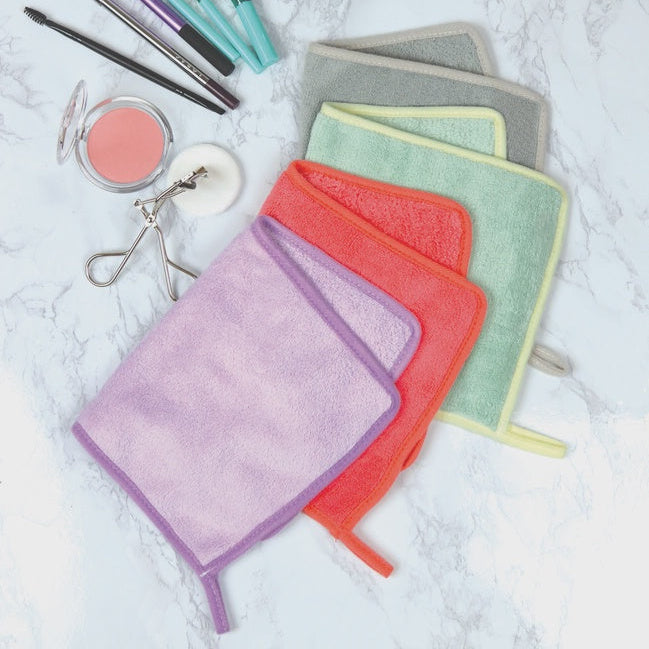 Photograph of colorful microfiber makeup removal cloths on a white marble surface featuring a silver eyelash curler, a peach blush compact, and vibrant purple, coral, and mint towels