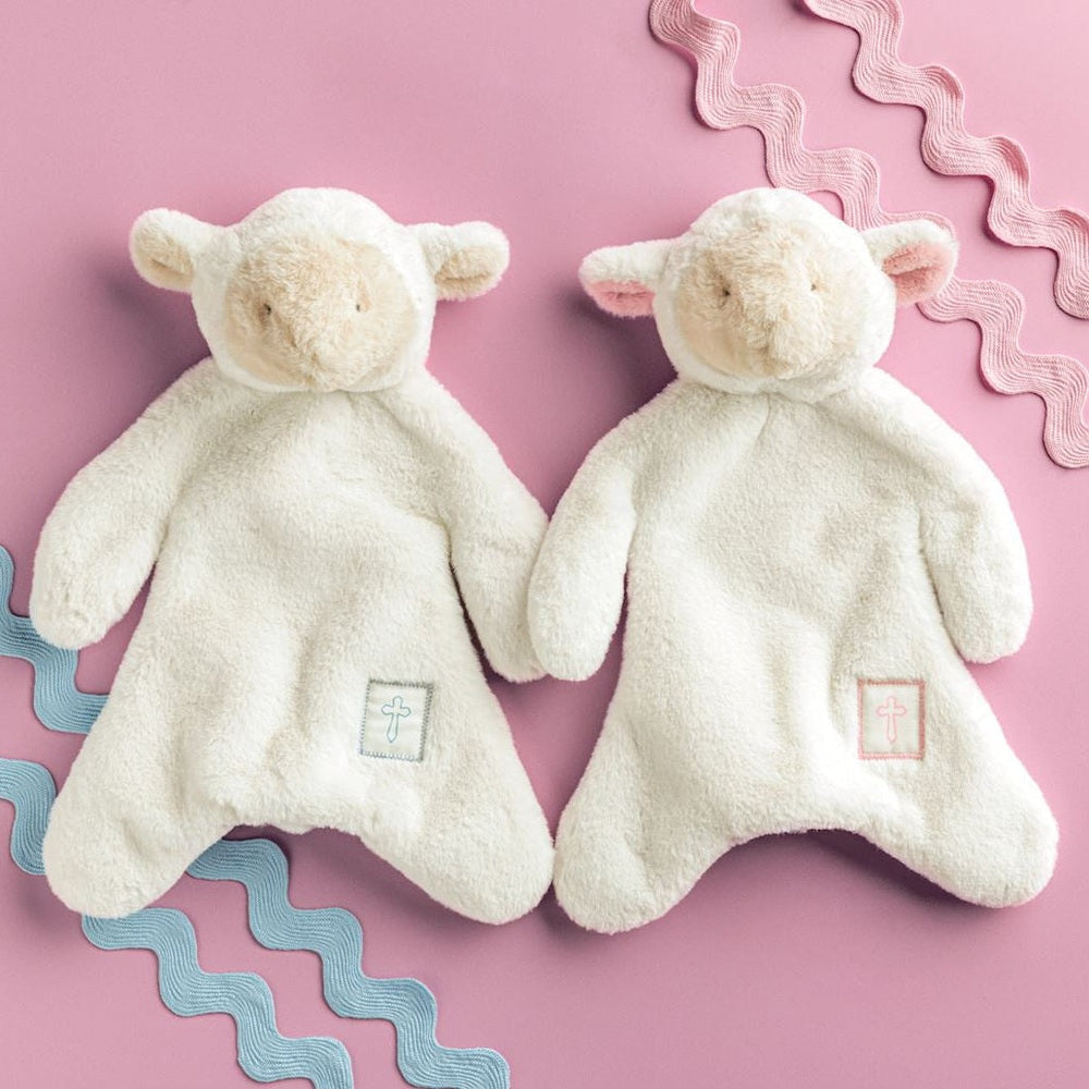 Studio photograph of two cream-colored plush lamb security blankets on a pink background featuring blue and pink wavy rickrack and small embroidered cross emblems