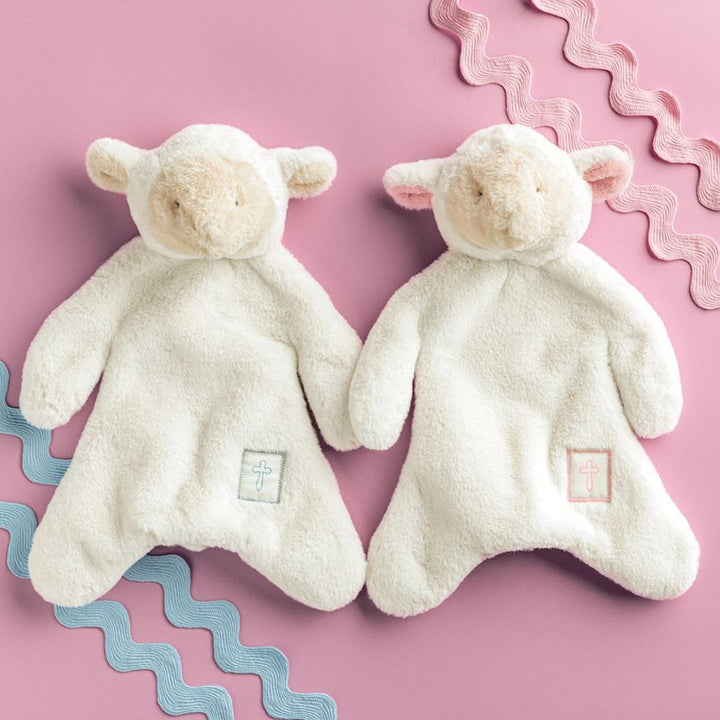 Studio photograph of two cream-colored plush lamb security blankets on a pink background featuring blue and pink wavy rickrack and small embroidered cross emblems