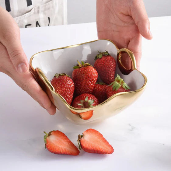 Ceramic gold-plated bowl held by hands over a white marble surface filled with vibrant red strawberries featuring unique wavy scalloped edges and gold-finished circular handles with two sliced fruit halves in the foreground