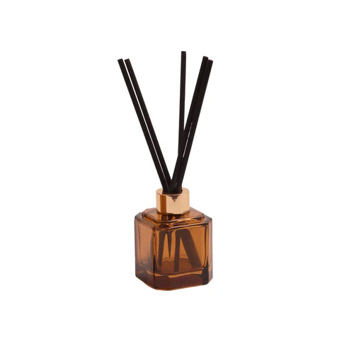 Commercial product photography of a square amber glass reed diffuser with a gold metallic collar and black fiber sticks centered against a minimalist white background