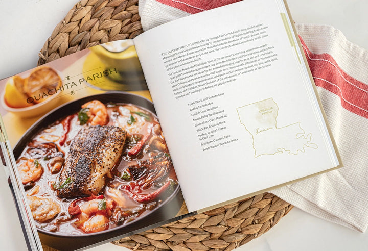 Photograph of an open cookbook highlighting the culinary heritage of Ouachita Parish, Louisiana, featuring a rich catfish courtbouillon with shrimp next to a gold map outline and a red-striped kitchen towel on a woven mat