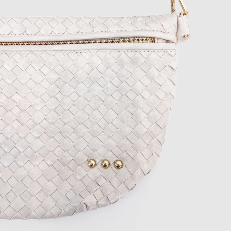 Photograph of a cream woven half-moon handbag on a white background featuring a gold-tone zipper, three decorative metallic studs, and an intricate braided texture
