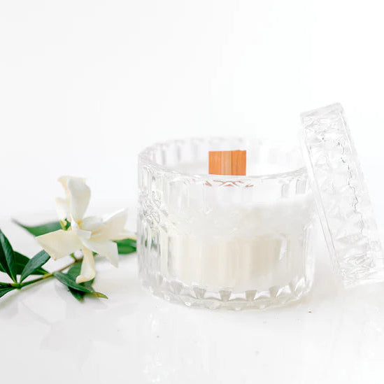 Photograph of a white scented candle featuring a wide wooden wick in a diamond-textured glass jar with its lid propped beside a single white gardenia flower on a minimalist white surface
