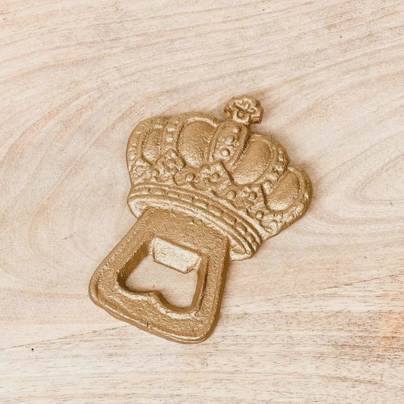 Cast iron crown bottle opener featuring a textured gold finish and intricate embossed details resting on a light-toned wooden surface