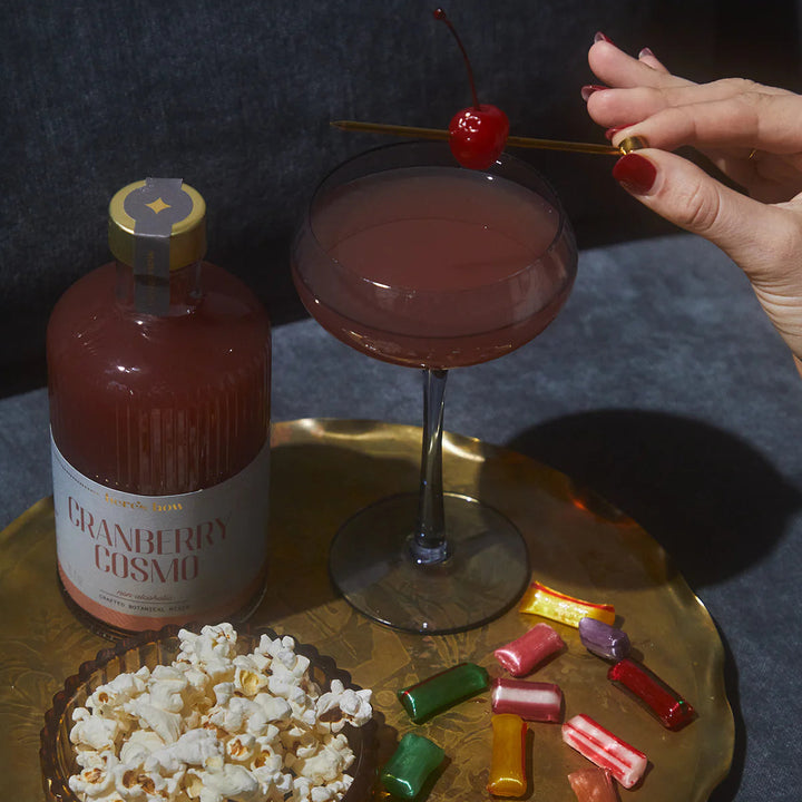 Hand placing a cherry on a gold pick over a pink cocktail beside a Cranberry Cosmo mixer, popcorn and candy