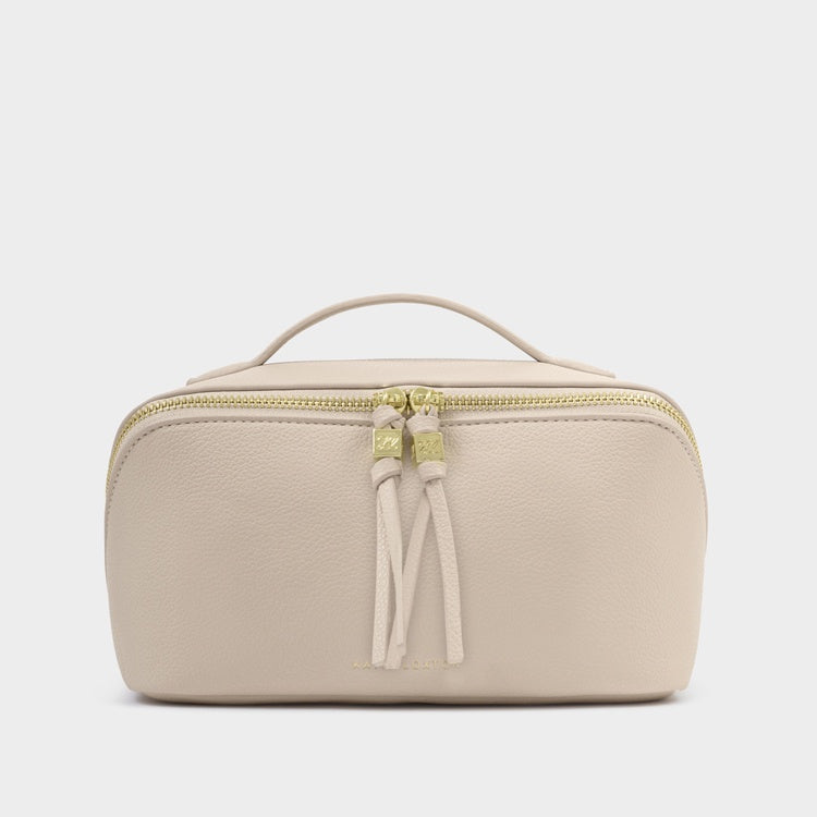 Pebbled vegan leather makeup bag by Katie Loxton featuring a soft beige finish, dual gold-toned zippers with elongated knotted tassels, and a structured top handle set against a minimalist gray background