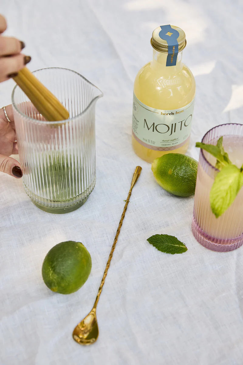 Hand muddling in a ribbed glass pitcher beside a Mojito mixer bottle, limes, mint and a gold bar spoon