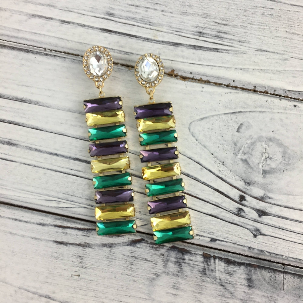 Jewelry earrings featuring long drops of tiered purple, yellow, and emerald green baguette crystals suspended from sparkling round rhinestone studs on a white distressed wooden surface