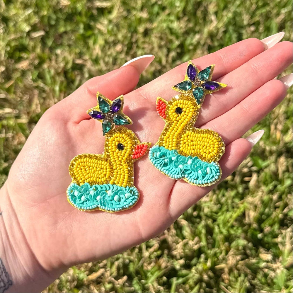 Handcrafted beaded and sequined earrings of yellow rubber ducks held in a palm over a green grass background featuring turquoise sequin water bases and vibrant jewel-toned rhinestone stars