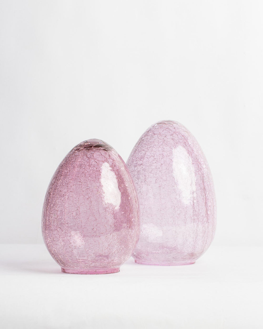 Hand-blown crackle glass sculptures of two translucent pink egg-shaped vessels positioned on a minimalist white studio surface featuring intricate internal webbing and glossy light reflections
