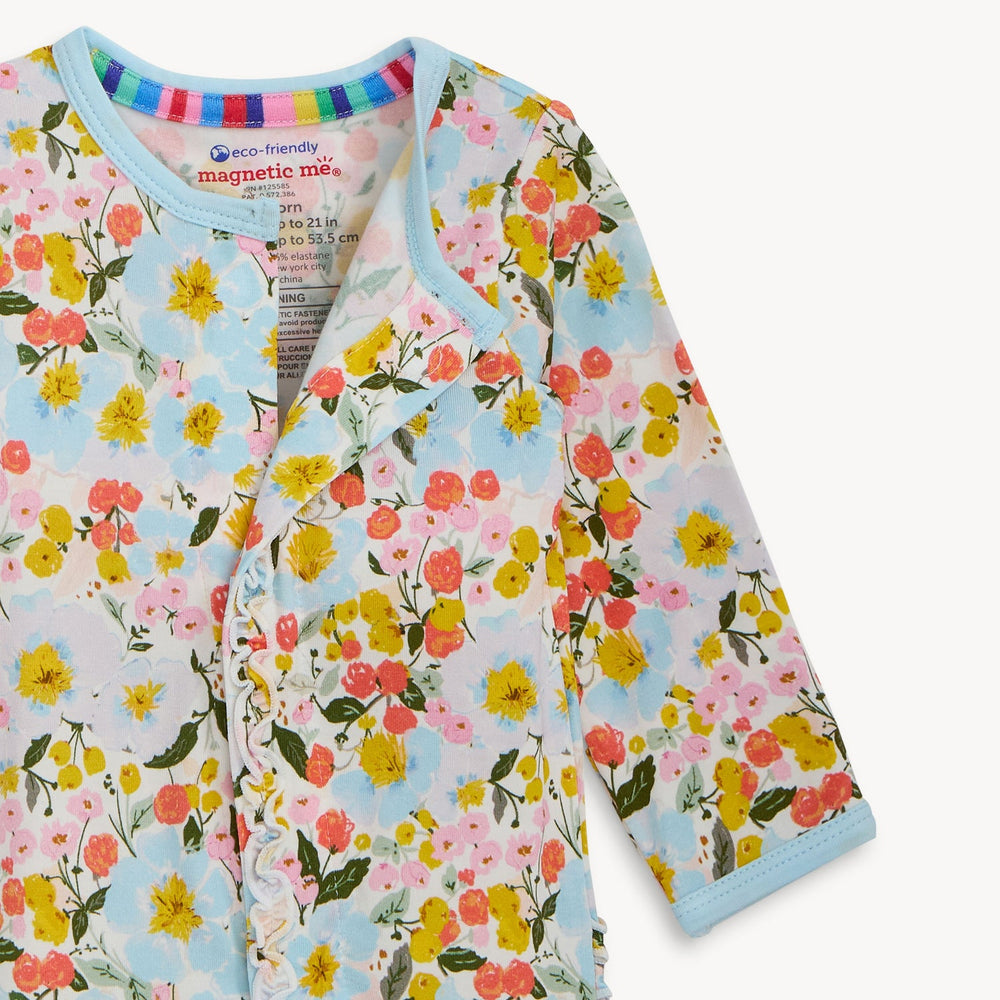 Photograph of a Magnetic Me floral baby onesie featuring a vibrant wildflower print with yellow and pink blossoms, light blue ruffled trim, and a rainbow-striped interior collar band