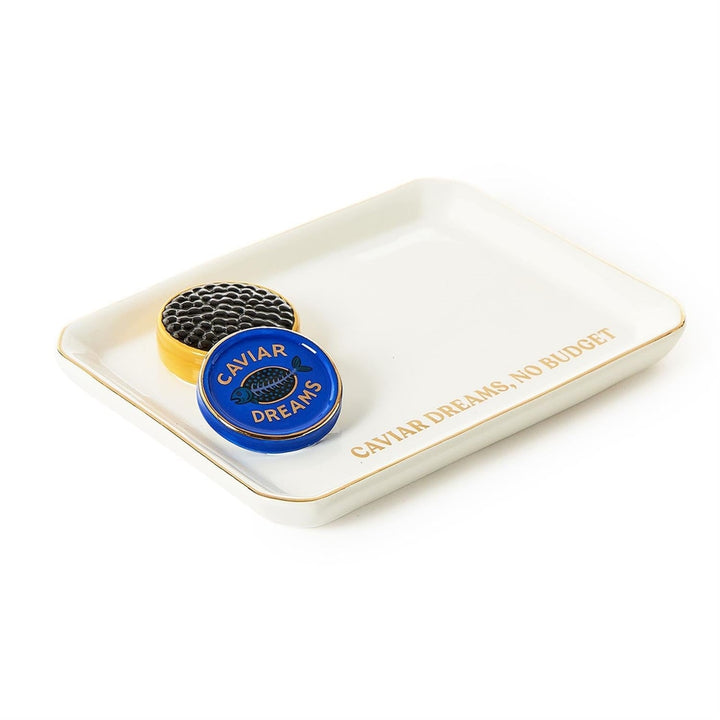 Porcelain trinket tray by Jonathan Adler featuring a sculptural miniature caviar tin with black beads and a blue lid next to gold-foil lettering that says CAVIAR DREAMS NO BUDGET on a white background with a metallic gold rim
