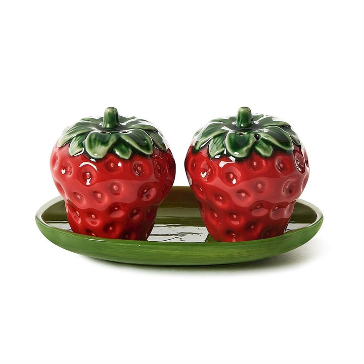 Ceramic strawberry salt and pepper shakers resting on a leaf-green tray featuring glossy red glazes, textured seed dimples, and sculpted green leaves against a neutral white background