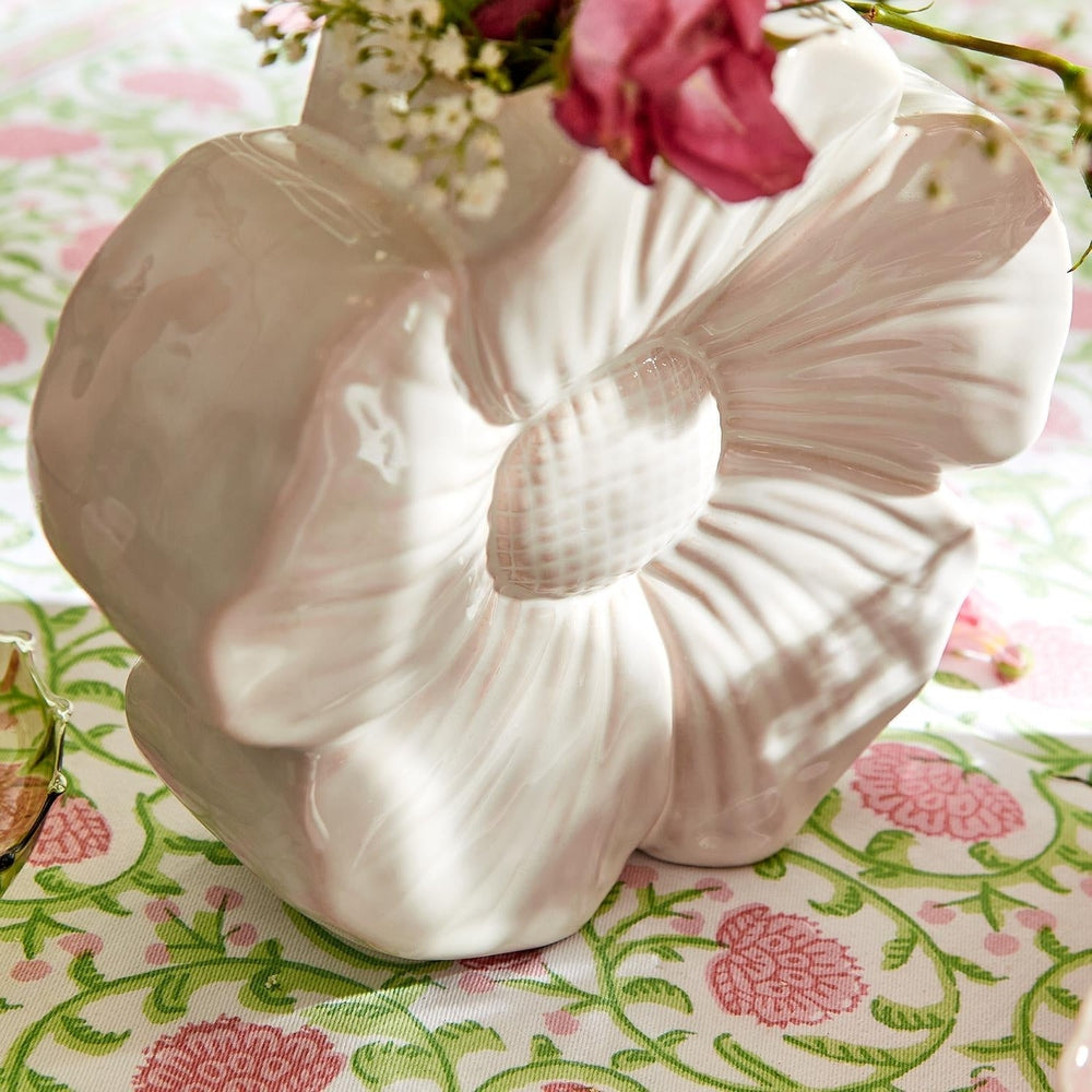 Ceramic flower-shaped vase by an unknown artist displayed on a pink and green floral tablecloth, highlighting its textured white petals, cross-hatched center, and vibrant pink flowers emerging from the top
