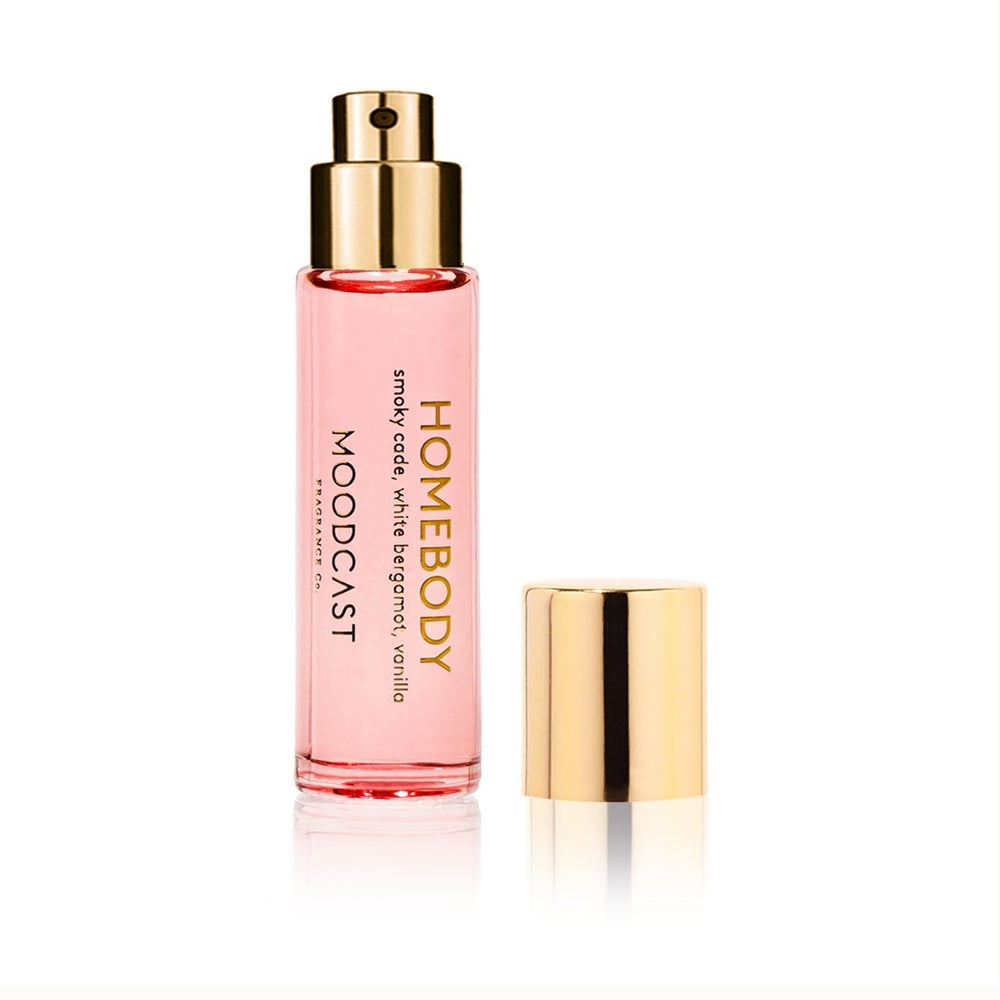 Product photography by Moodcast Fragrance Co of a Homebody perfume spray featuring a slender glass vial of blush pink liquid with a metallic gold-tone atomizer and a detached gold cap set against a reflective white background
