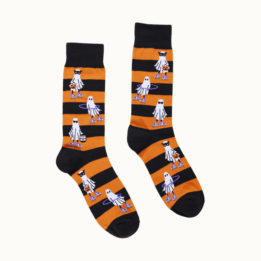 A pair of patterned crew socks displayed on a solid green background featuring vibrant orange and black stripes with whimsical ghosts on purple roller skates wearing black sunglasses and carrying orange pumpkin pails