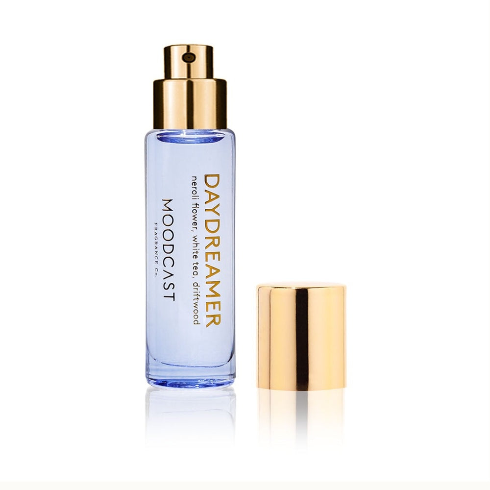 Studio product photograph by Moodcast Fragrance Co of a Daydreamer perfume bottle on a white reflective background featuring pale lavender liquid, gold-foiled labels for neroli flower and white tea, and a polished metallic gold spray nozzle with a detached cap