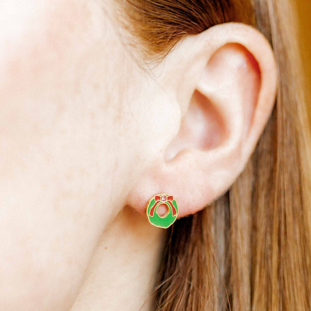 Photograph of a person's ear featuring a vibrant green enamel Christmas wreath earring adorned with a tiny red bow, a sparkling central jewel, and gold-finished edges