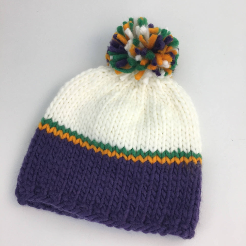 Hand-knitted yarn beanie by an unknown artisan featuring a chunky purple base and off-white top separated by thin orange and green stripes with a multi-colored pom-pom on a plain background