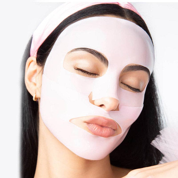 Commercial photograph of a woman wearing a translucent light pink sheet mask in a studio setting featuring a matching pink headband and a small gold hoop earring