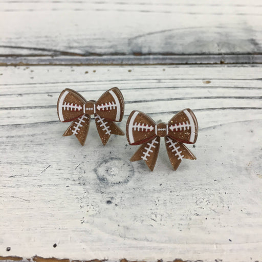Acrylic resin stud earrings featuring brown football-themed bows with white laces and gold glitter accents are displayed on a rustic white wooden tabletop
