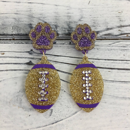 Handmade resin dangle earrings featuring purple and gold glitter paw prints and footballs with sparkling rhinestone laces displayed on a white-washed wood surface
