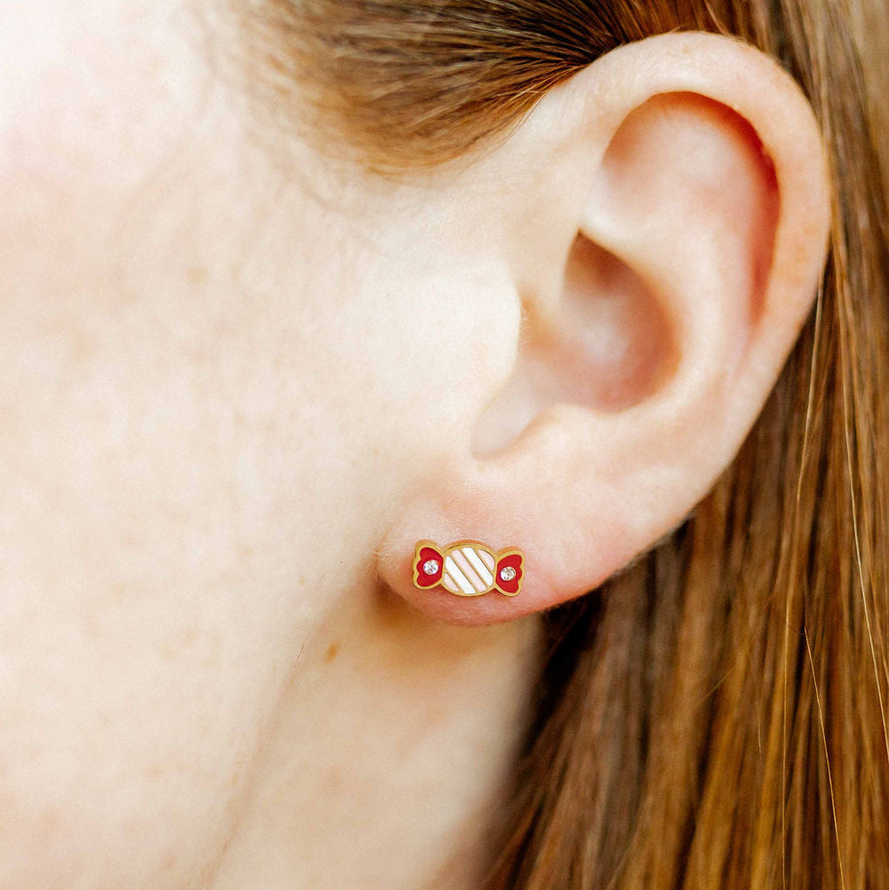 Photograph of a gold candy-shaped stud earring worn on a person's ear featuring a red and white striped enamel body and shimmering crystal accents on the wrapper ends